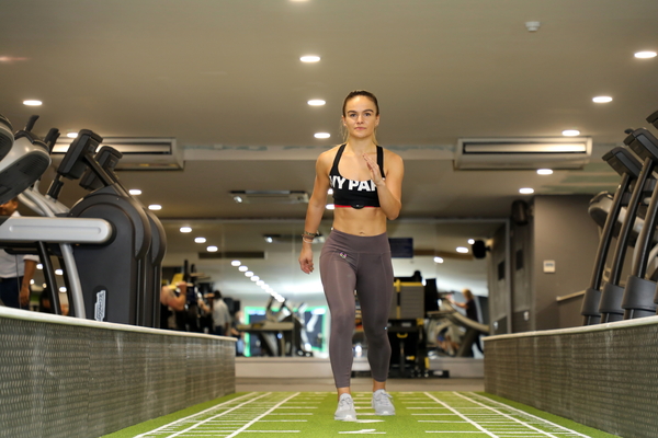 A woman in sportswear runs towards the camera along a green artificial turf track in a gym, exercise machines either side of her
