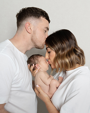 A mother and father in white shirts each kiss the head of their newborn baby, held between them against a pale wall